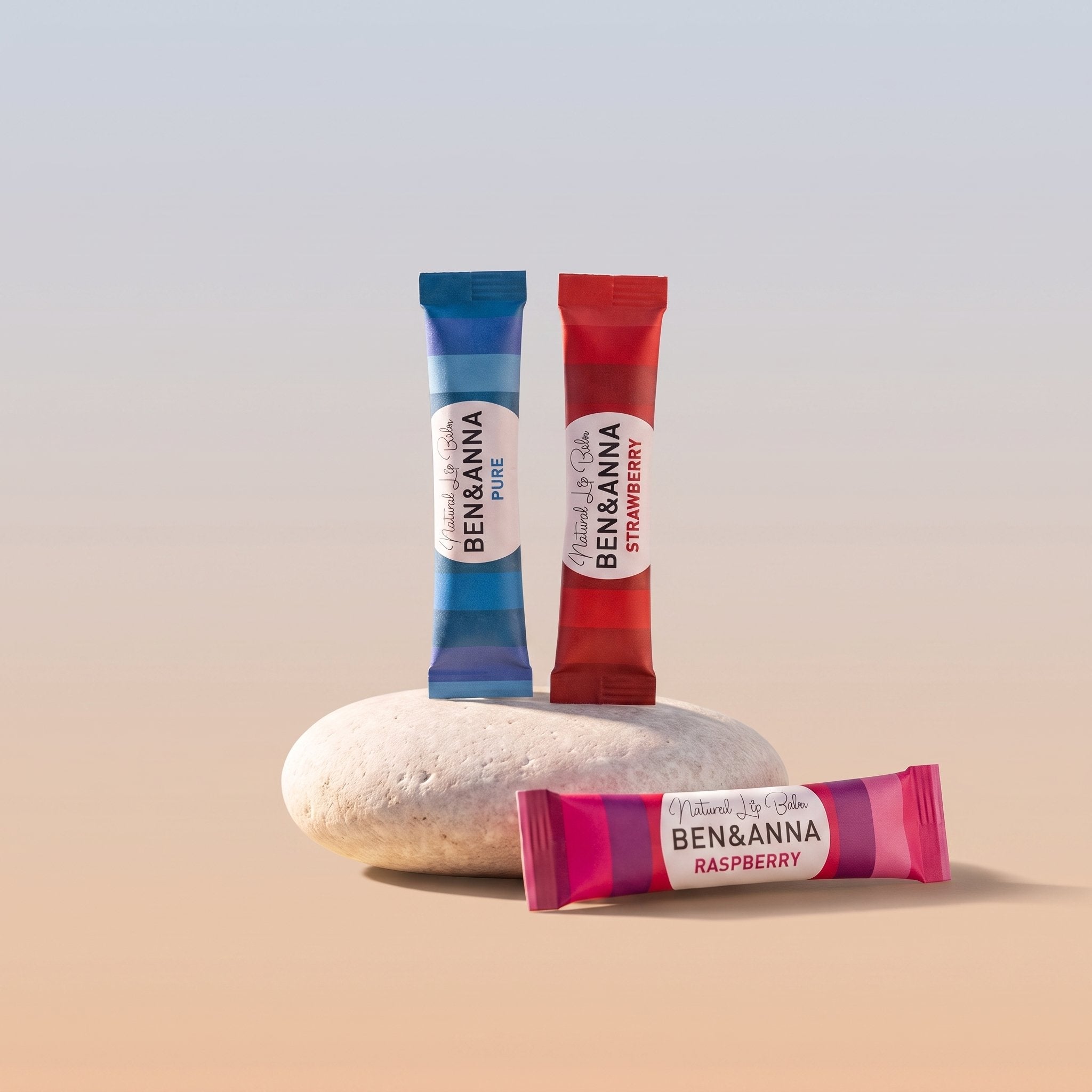 Drei BEN&ANNA Natural Lip Balm Papertuben in Pure, Strawberry und Raspberry auf einem hellen Stein vor sanftem Farbverlauf [KI]