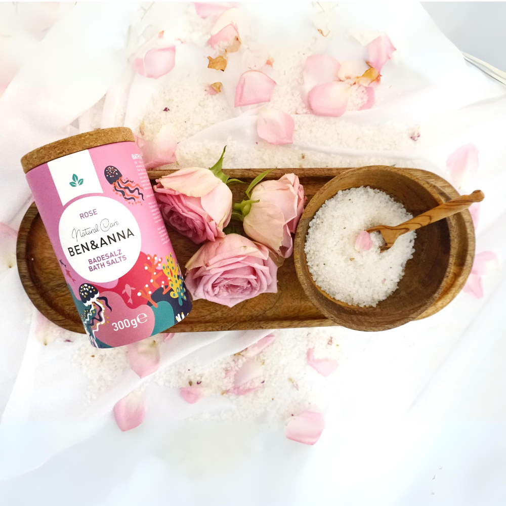 Badesalz mit Rosenduft von BEN&ANNA, in einer weißen Schale mit rosa Rosenblüten und Duftkerzen, für entspannendes Bad.